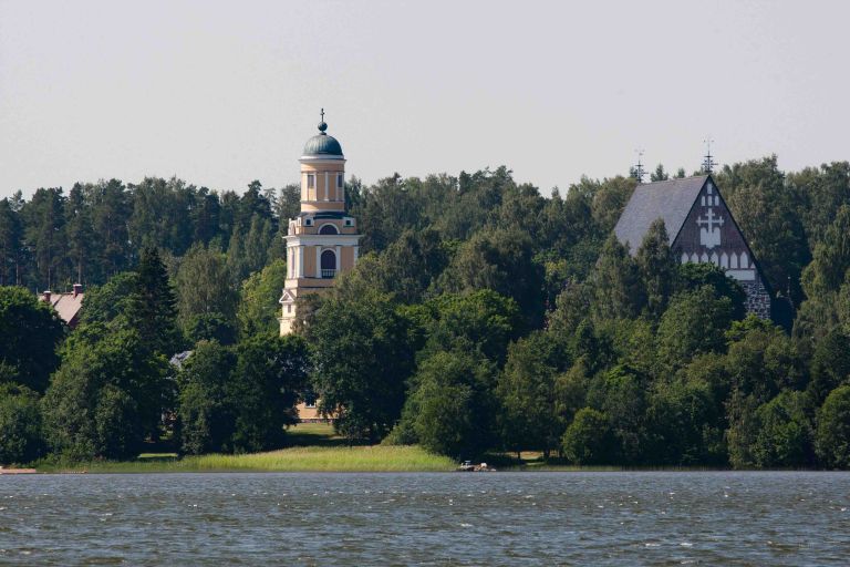 Maisema, jossa veden äärellä puiden keskeltä erottuu vanha kivinen keskiaikainen kirkko.